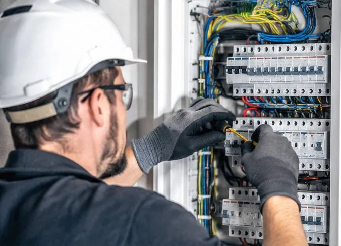 An electrician in a hard hat and safety glasses working inside an open consumer unit, tightening a connection on one breaker in a row of them.