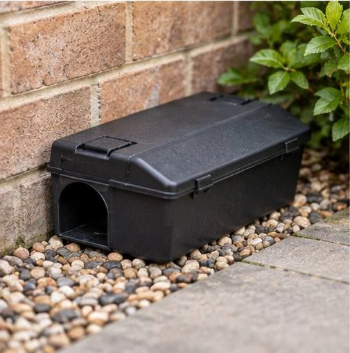 A black tamper-resistant bait box sitting tight against a brick wall on a gravel border, its entry tunnel facing along the run of the wall.